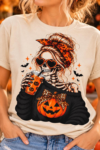 A woman wearing a sand-colored t-shirt featuring a graphic of a skeletal woman with heart sunglasses and a pumpkin.