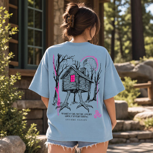 May include: A light blue t-shirt featuring a black ink illustration of a cabin on chicken legs in a forest under a moon. The cabin window is bright pink. Text reads: WOMEN? OH, WE'RE ANGRY ABOUT EVERYTHING. ENCORE NATION. The design includes pink accents.
