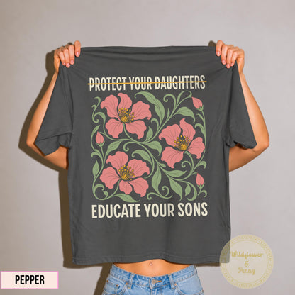 May include: A charcoal gray t-shirt featuring the text PROTECT YOUR DAUGHTERS with a line through it and EDUCATE YOUR SONS in cream lettering. The center graphic displays three large pink flowers with yellow centers surrounded by green leafy vines.