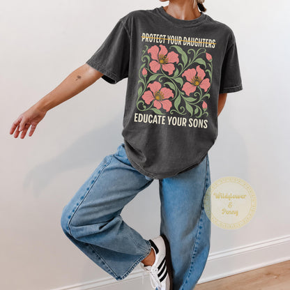 May include: A charcoal gray short-sleeve t-shirt featuring a graphic print of pink flowers with green leaves. The text reads PROTECT YOUR DAUGHTERS with a line through it and EDUCATE YOUR SONS below the floral design in a cream-colored font.