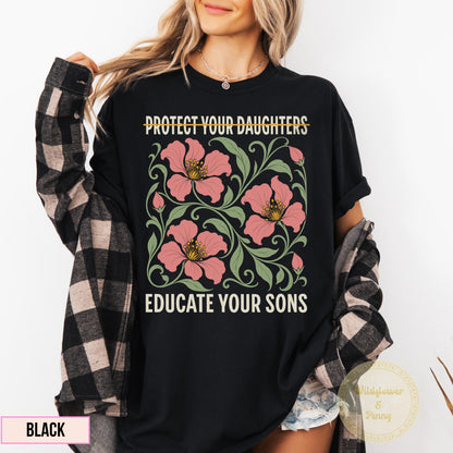 May include: A black t-shirt featuring a graphic print with the text PROTECT YOUR DAUGHTERS at the top and EDUCATE YOUR SONS at the bottom. The center design displays pink flowers with green leaves and swirling vines on a dark background.