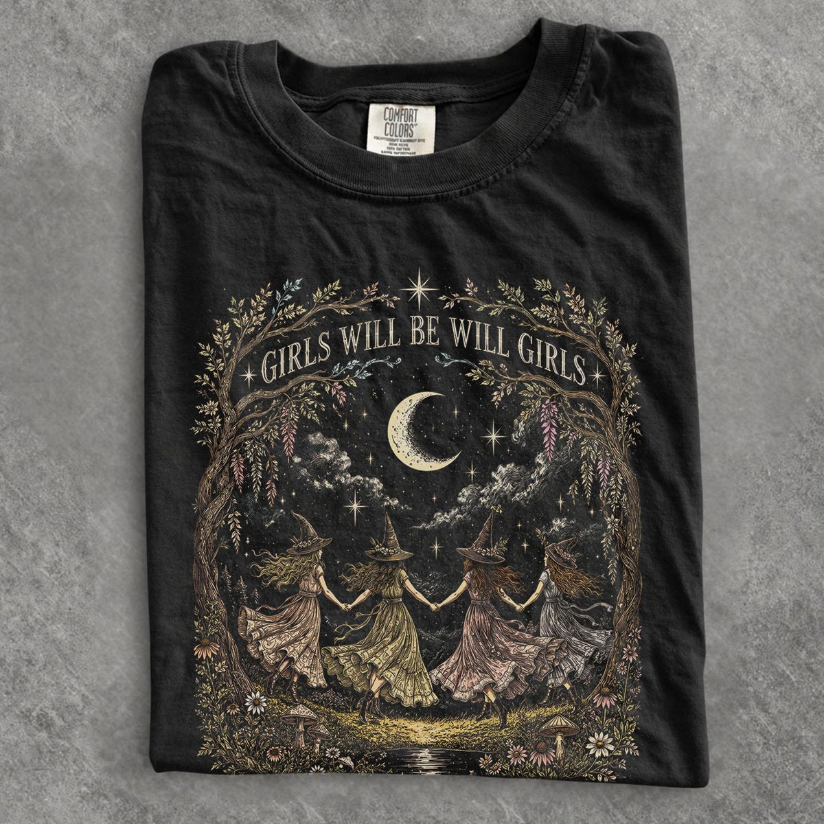 May include: A black t-shirt featuring a graphic print of four figures in pointed hats holding hands in a circle under a crescent moon and stars. The design is framed by trees and flowers with the text GIRLS WILL BE WILL GIRLS arched above.