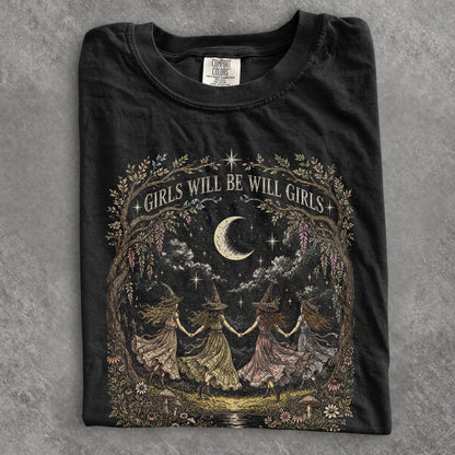 May include: A black t-shirt featuring a graphic print of four figures in pointed hats holding hands in a circle under a crescent moon and stars. The design is framed by trees and flowers with the text GIRLS WILL BE WILL GIRLS arched above.