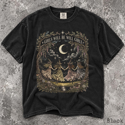 May include: A black short-sleeve t-shirt featuring a graphic print of four figures in pointed hats holding hands in a circle under a crescent moon. The text "GIRLS WILL BE WILL GIRLS" is arched above the figures, surrounded by trees and flowers.
