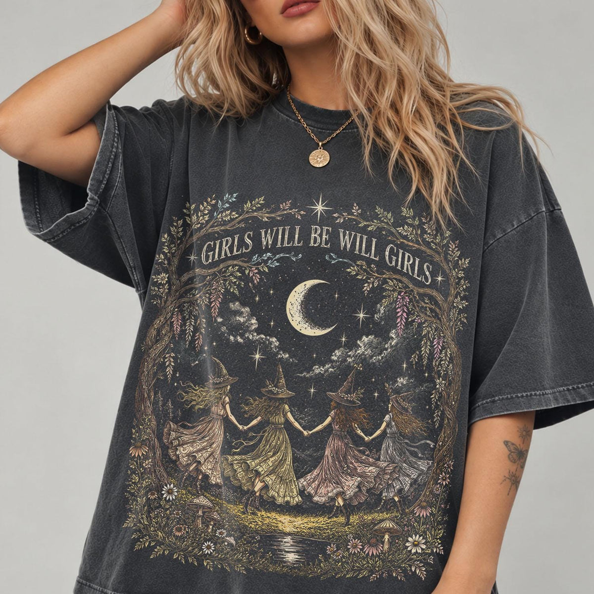 May include: A charcoal gray oversized t-shirt featuring a graphic print of four figures in pointed hats holding hands in a circle under a crescent moon. The design includes floral arches, stars, and the text GIRLS WILL BE WILL GIRLS in white.