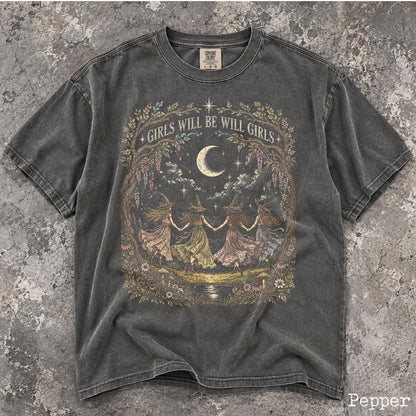 May include: A charcoal gray short-sleeve t-shirt featuring a graphic print of four figures in pointed hats holding hands under a crescent moon. The text "GIRLS WILL BE WILL GIRLS" is arched above the figures, surrounded by a detailed floral border.