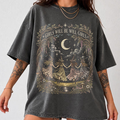 May include: A charcoal gray short-sleeve t-shirt featuring a graphic print of four figures in pointed hats holding hands in a circle under a crescent moon. The text GIRLS WILL BE WILL GIRLS is printed above the illustration, surrounded by foliage.