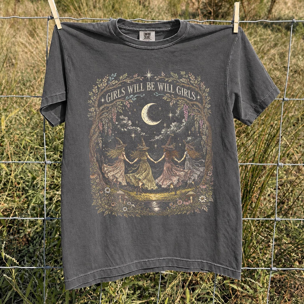 May include: A charcoal gray short-sleeve t-shirt featuring a graphic print of four figures in pointed hats holding hands in a circle under a crescent moon. The text "GIRLS WILL BE WILL GIRLS" is printed above the illustration, surrounded by floral details.