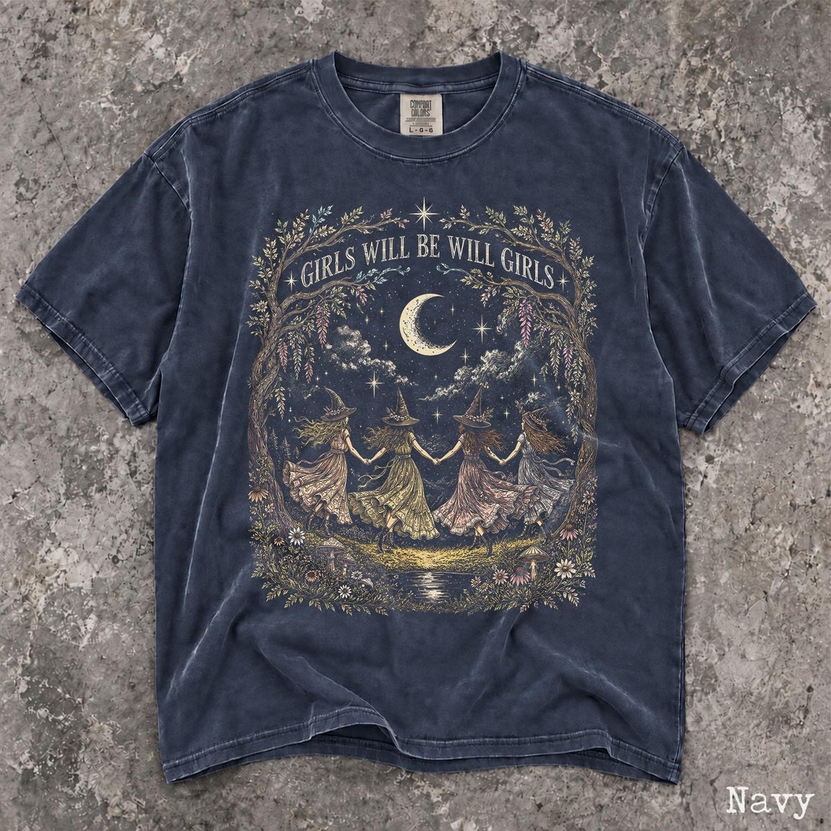 May include: A navy blue short-sleeve t-shirt featuring a graphic print of four figures in pointed hats holding hands in a circle under a crescent moon. The text "GIRLS WILL BE WILL GIRLS" is arched above the scene, which is framed by floral vines.