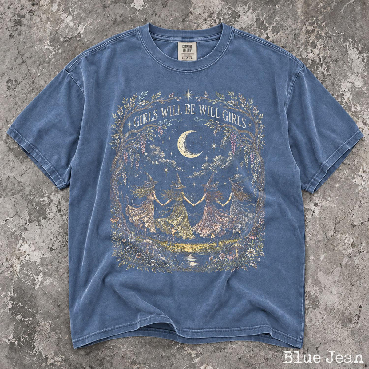 May include: A blue jean colored short-sleeve t-shirt featuring a graphic print of four figures in pointed hats dancing in a circle under a crescent moon. The text GIRLS WILL BE WILL GIRLS is arched above the scene, framed by floral and vine illustrations.