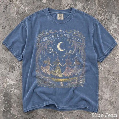May include: A blue jean colored short-sleeve t-shirt featuring a graphic print of four figures in pointed hats dancing in a circle under a crescent moon. The text GIRLS WILL BE WILL GIRLS is arched above the scene, framed by floral and vine illustrations.