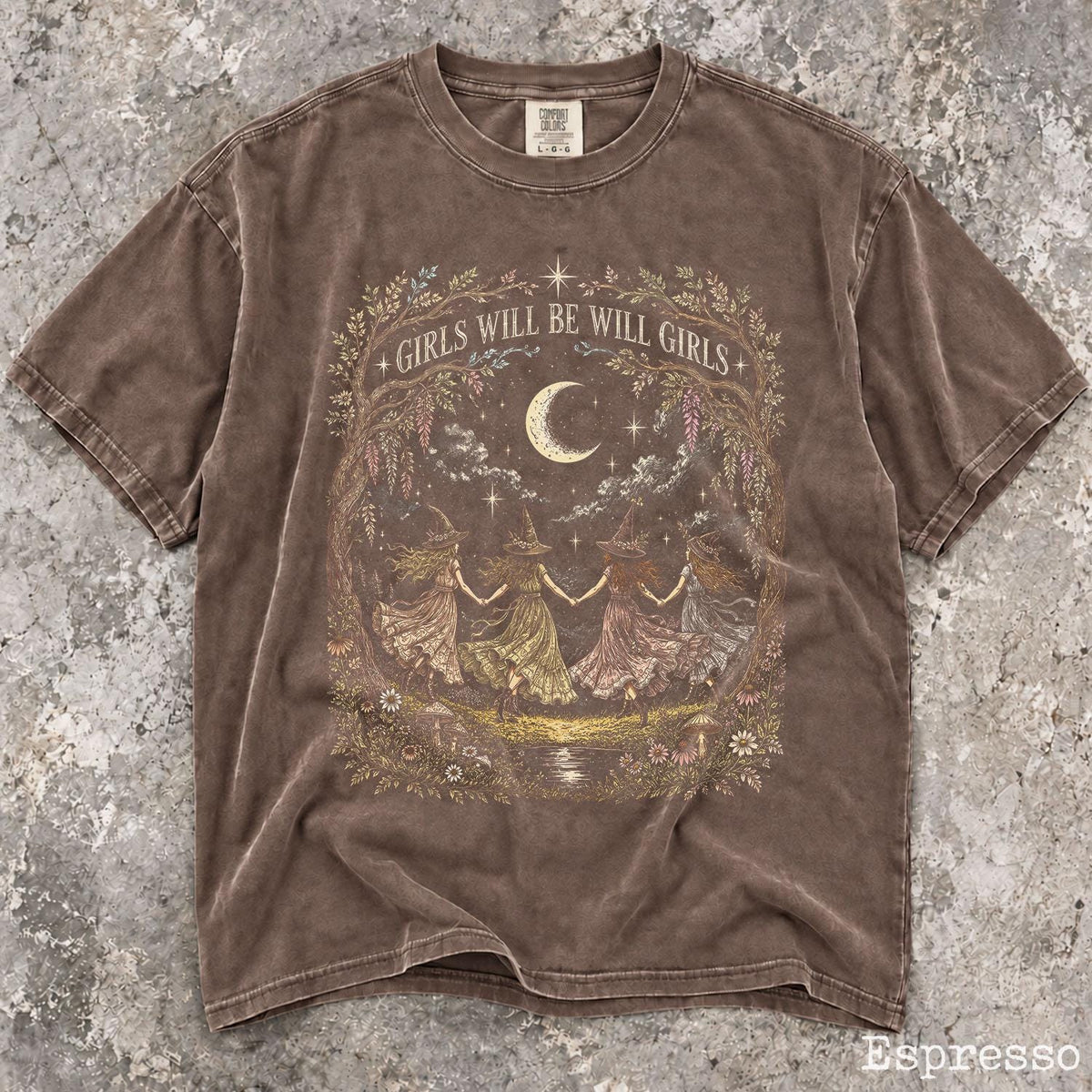 May include: A brown short-sleeve t-shirt featuring a graphic print of four figures in hats holding hands in a circle under a crescent moon. The text GIRLS WILL BE WILL GIRLS is printed above the moon. The design is framed by a floral wreath.
