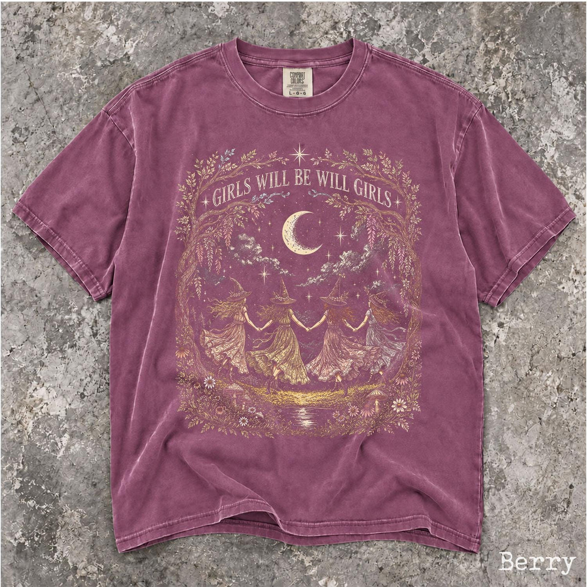 May include: A berry-colored short-sleeve t-shirt featuring a gold graphic print of four figures in pointed hats holding hands in a circle under a crescent moon. The text GIRLS WILL BE WILL GIRLS is printed in an arch above the central illustration.