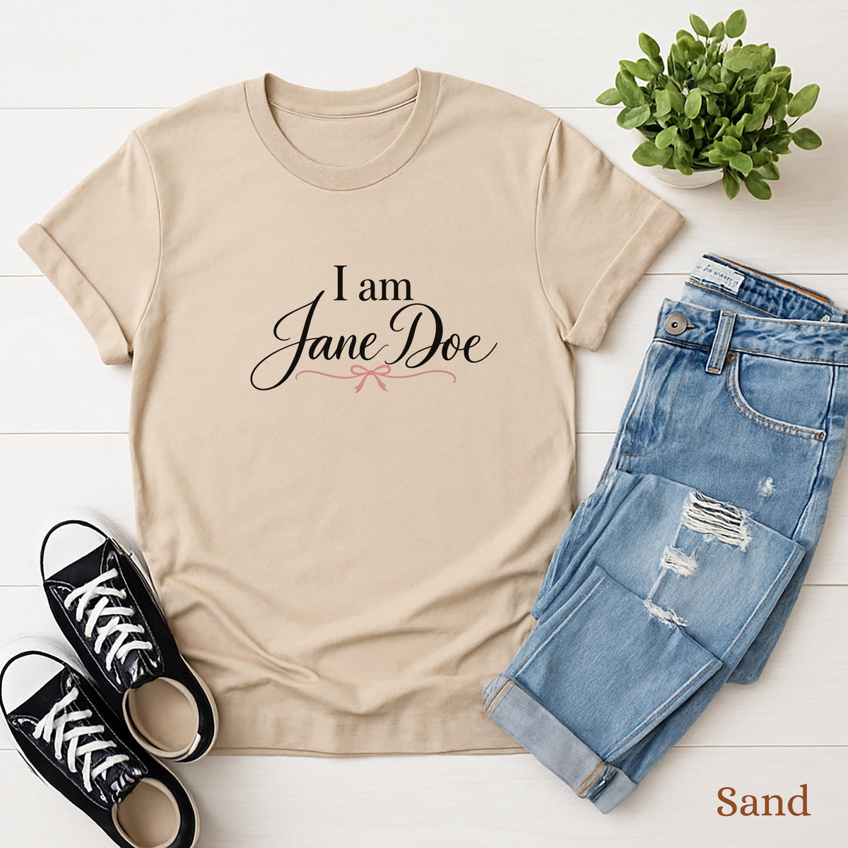 A beige t-shirt with the text "I am Jane Doe" printed on it, along with a pair of blue jeans and black sneakers, all laid out on a white background.