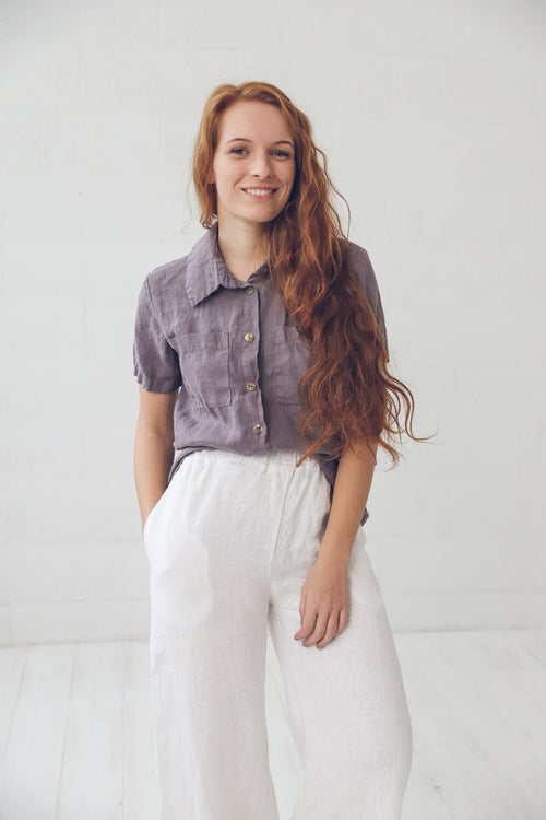 May include: A woman wearing a purple linen shirt with a white linen button-down collar and white linen pants.
