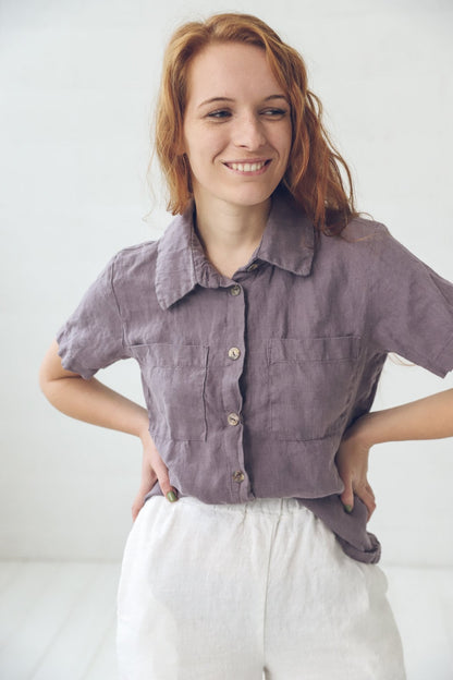May include: A woman wearing a purple linen shirt with a white linen skirt. The shirt has a collar and short sleeves. The skirt has a high waist and is loose fitting.