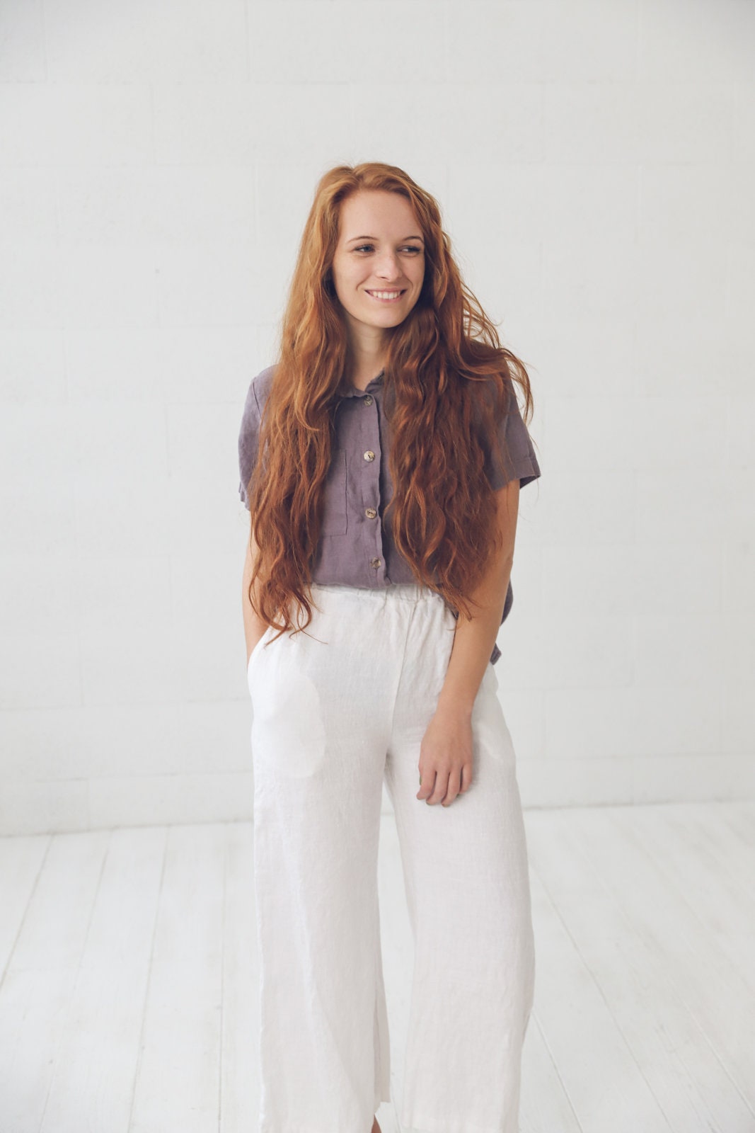 May include: A woman with long, red hair wearing a purple linen shirt and white linen pants.