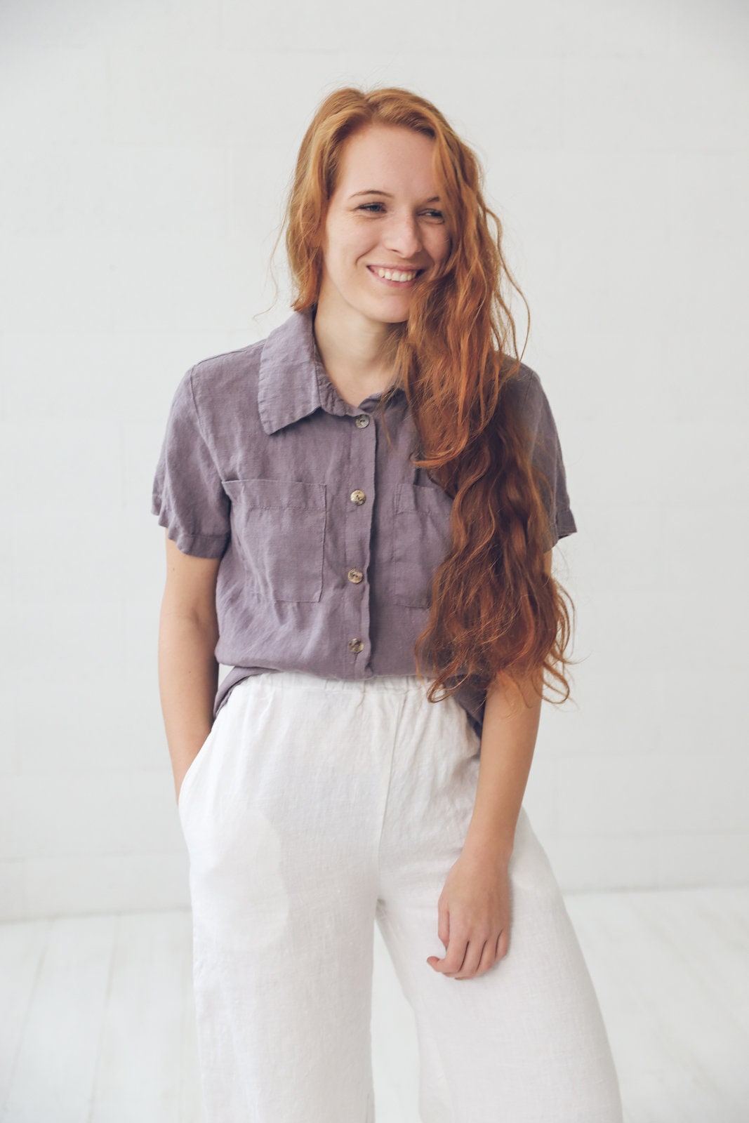 May include: A woman wearing a purple linen shirt with a white linen pants. She is smiling and looking to the side.