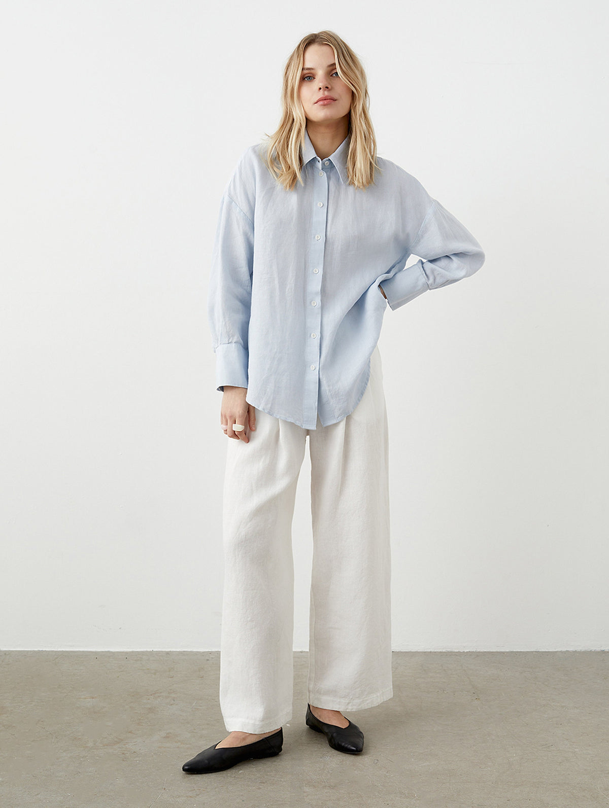 May include: A woman wearing a light blue button-down shirt and white wide-leg pants. The shirt has long sleeves and a classic collar. The pants are loose-fitting and made of a light-colored fabric. Black flats complete the outfit, creating a casual, yet stylish look.