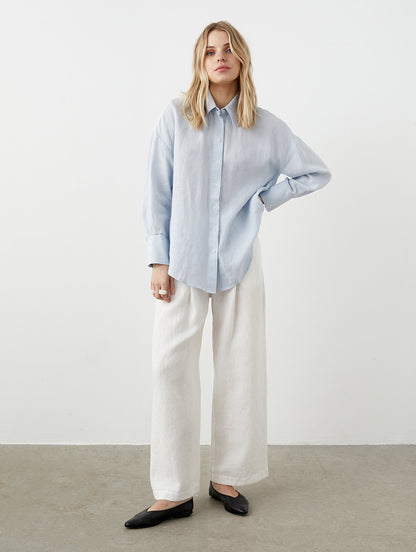 May include: A woman wearing a light blue button-down shirt and white wide-leg pants. The shirt has long sleeves and a classic collar. The pants are loose-fitting and made of a light-colored fabric. Black flats complete the outfit, creating a casual, yet stylish look.
