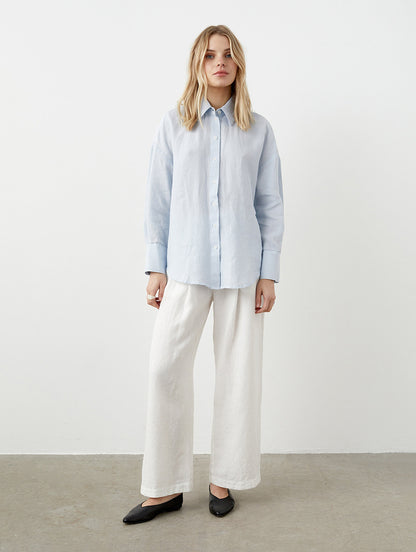 May include: A light blue button-down shirt and white wide-leg pants. The long-sleeved shirt has a classic collar and cuffs. The pants are a loose fit. Black flats complete the outfit, creating a minimalist, casual style.