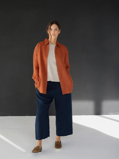 May include: A woman wearing a rust-colored linen shirt over a light-colored top, paired with navy blue culottes. The outfit is completed with brown loafers. The background is a dark gray wall and a white floor.