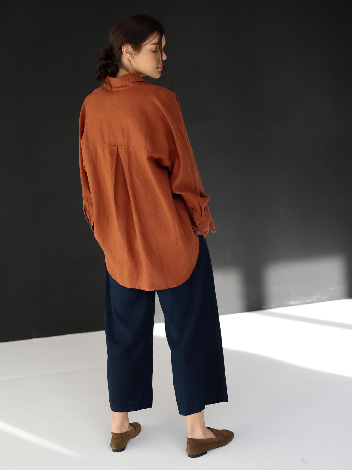 May include: A rust-colored linen shirt with long sleeves and a button-down front is paired with navy blue wide-leg pants. The outfit is completed with brown suede loafers. The clothing is casual and comfortable.