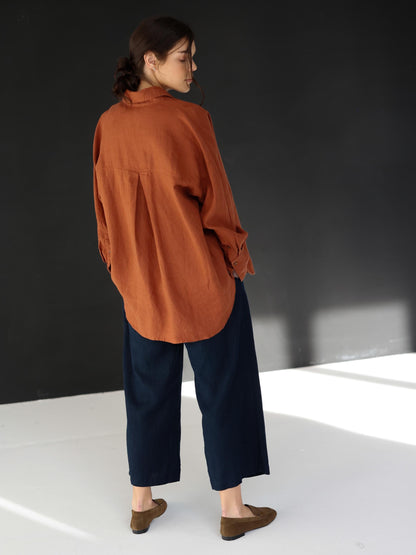 May include: A rust-colored linen shirt with long sleeves and a button-down front is paired with navy blue wide-leg pants. The outfit is completed with brown suede loafers. The clothing is casual and comfortable.
