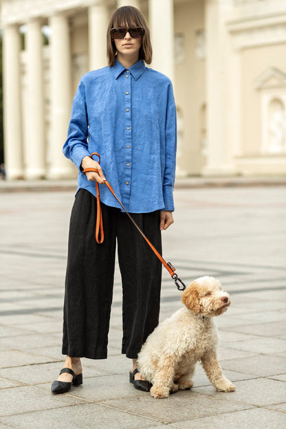 May include: A person wearing a blue button-down shirt and black pants, holding an orange dog leash attached to a small, cream-colored dog. The dog is sitting on a paved surface.