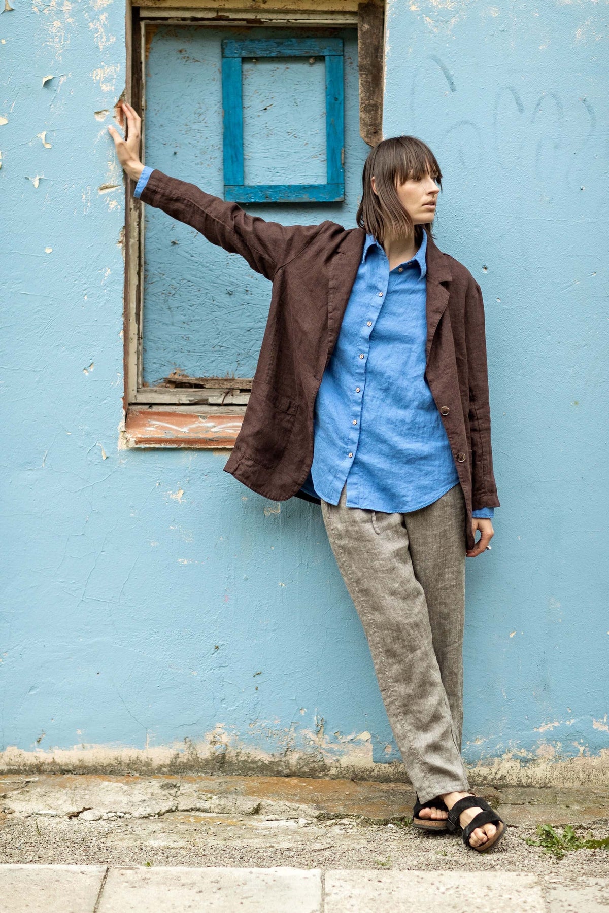 May include: A person wearing a brown linen blazer over a blue button-down shirt and gray linen pants. The person is leaning against a weathered blue wall. Black sandals complete the outfit.