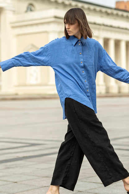 May include: A person wearing a blue linen button-down shirt and black linen pants. The shirt has a classic collar and wooden buttons. The pants are wide-legged. The outfit is casual and comfortable, suitable for everyday wear.